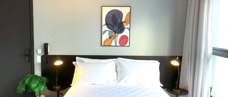 A neatly made bed with white linens, a potted plant, and a painting on the wall.