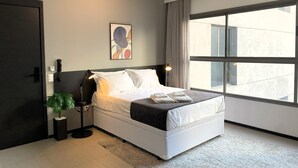 A modern bedroom with a large window, a bedside lamp, a plant, and a framed artwork on the wall. - Moderno com AC academia 24h e rooftop (São Paulo)