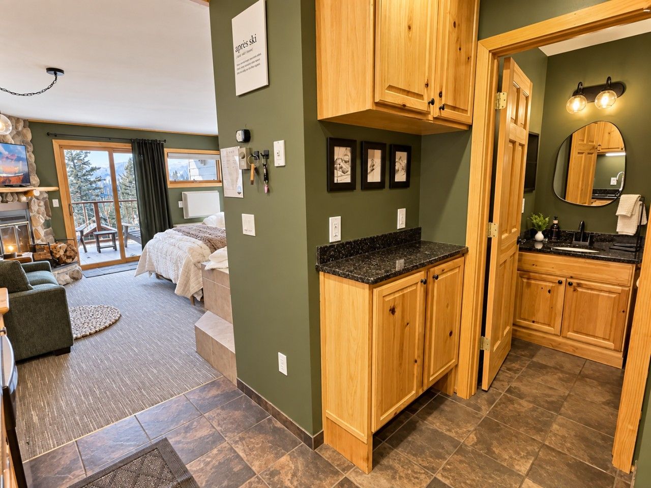 A room with a bed, a sitting area with a chair, a kitchen with wooden cabinets, and a large window.