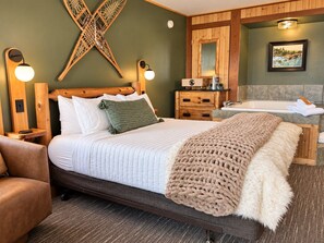 A hotel room with a bed, a wooden headboard, a bedside table, a lamp, a bench, and a bathroom with a bathtub and a towel rack. - Slope-Side Pet-Friendly Studio | In-Unit Jet Tub (Lutsen)