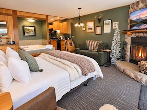 A cozy room with a bed, a fireplace, a stone fireplace surround, and a kitchen area.