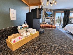 A kitchen with a coffee maker, a wooden tray with coffee cups, and a view of a bed and mountains through a window.