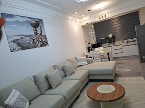 Ein modernes Wohnzimmer mit einem Ecksofa, einem Couchtisch und einer Küchenzeile im Hintergrund.