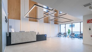 A modern reception area with a concrete reception desk, wooden accents, and a large window offering a view of the outside. - Micampus Foronda 7 (Madrid)