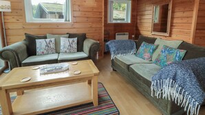 A cozy cabin interior with two sofas, a wooden table, and large windows offering views of the outdoors.