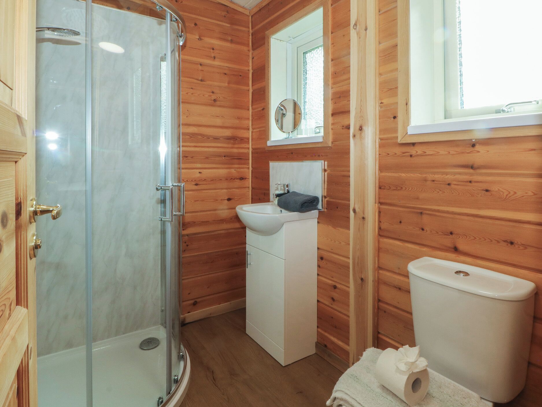 A wooden cabin bathroom with a glass shower enclosure, a sink, a toilet, and a mirror above the sink.