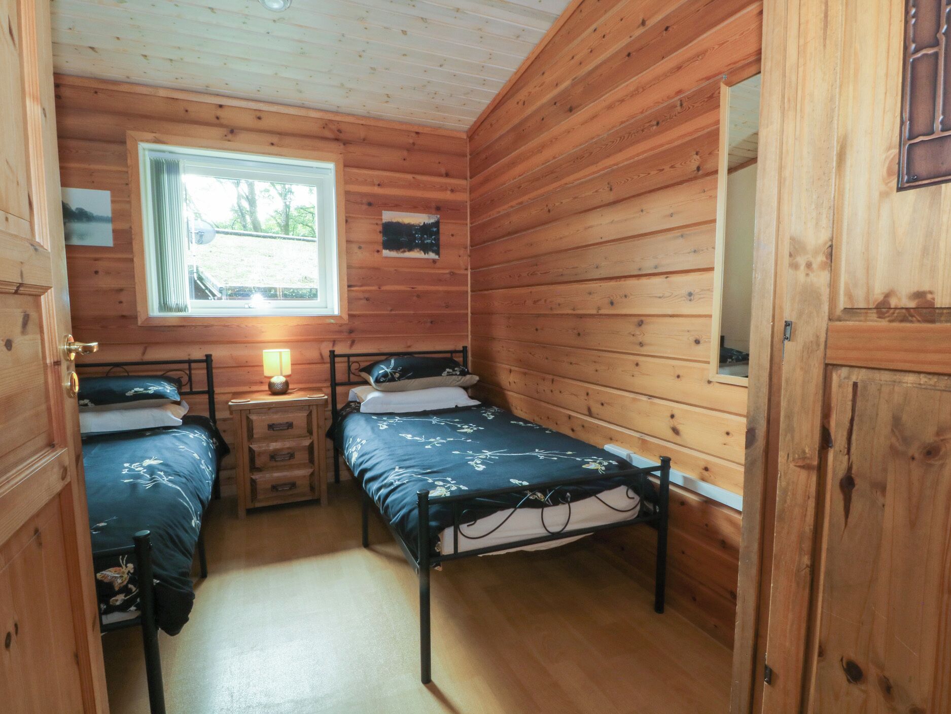 Une petite pièce agréable, aux murs en bois, avec deux lits simples. Une fenêtre laisse entrer la lumière naturelle, et une petite table avec une lampe est également présente.
