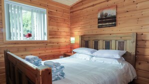 A wooden cabin bedroom with a bed, a nightstand, a window with blinds, and a painting on the wall.