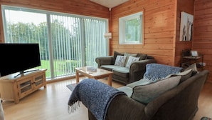 A cozy living room with a flat-screen TV, a sofa, a coffee table, and a large window with blinds.