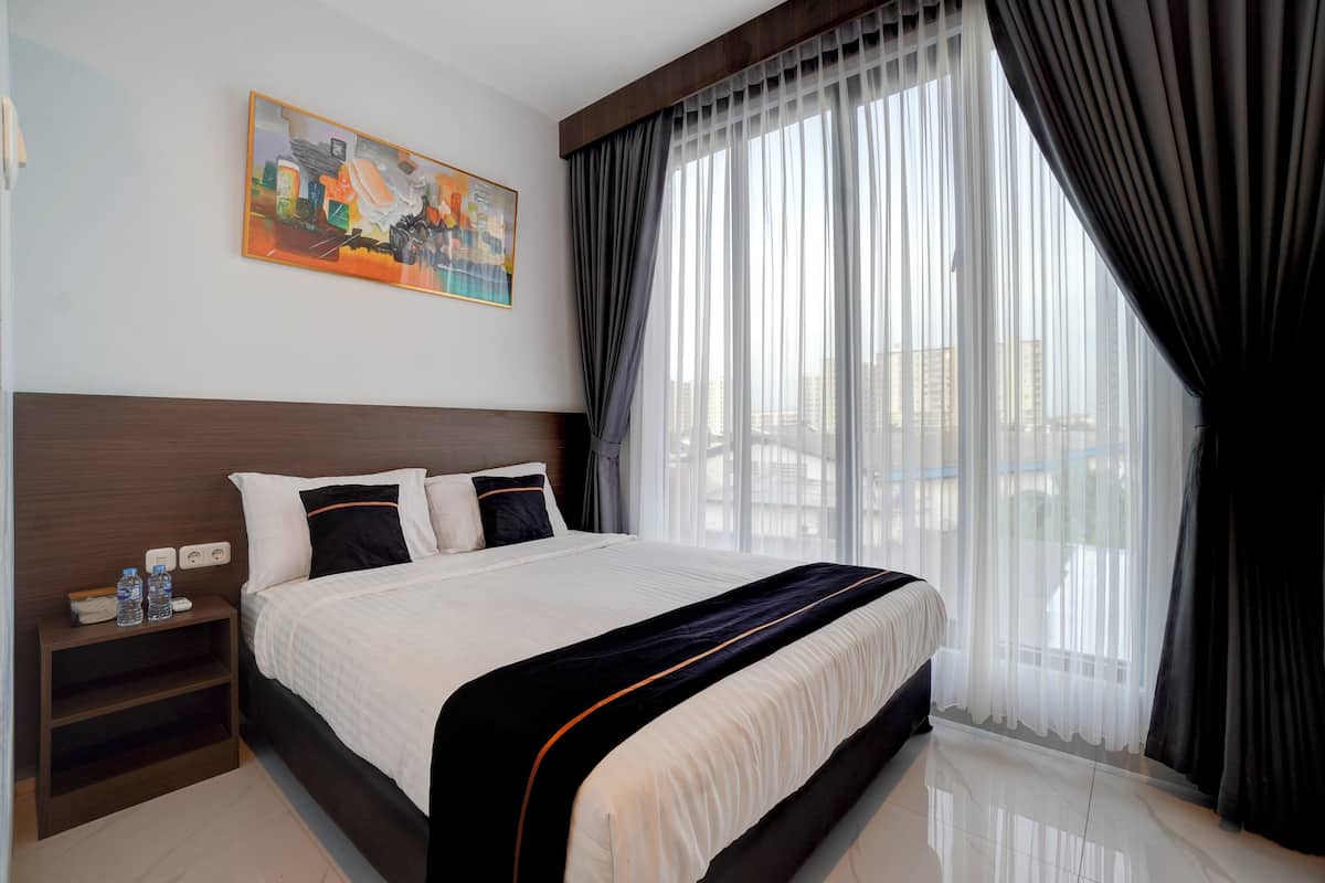 Quarto de hotel com uma cama grande, uma mesa de cabeceira com abajur, um quadro na parede e vista para edifícios através de cortinas translúcidas.