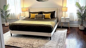 Un dormitorio con cama, dos mesitas de noche, ventilador de techo y dos ventanas con cortinas.