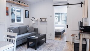 Una sala de estar moderna y bien iluminada con un sofá gris, una mesa de centro negra y una pequeña mesa blanca. Se ve una cama al fondo con una ventana y persianas, y una estantería con plantas.