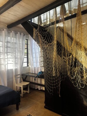 A room with a wooden ceiling, a black wall, and a window with sheer curtains. There is a chair and a table with a blue bag on it. - Refúgio Do Sagrado Morere  (Morere Boipeba)