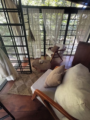 A room with a wooden floor, a cushioned chair, a small round table, and a window with a view of greenery. - Refúgio Do Sagrado Morere  (Morere Boipeba)