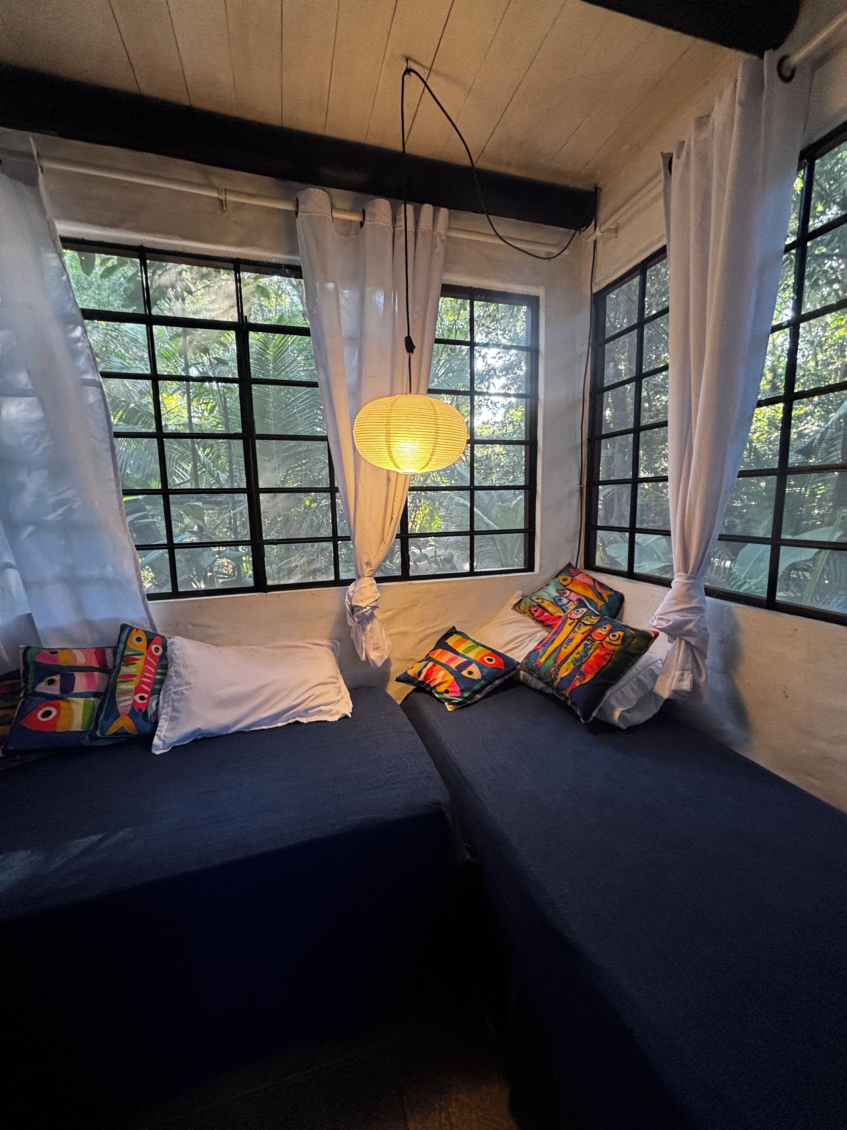 A room with two beds, a hanging light fixture, and a view of greenery through large windows.