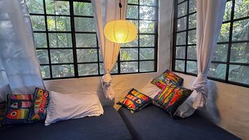 A room with two beds, a hanging light fixture, and a view of greenery through large windows.