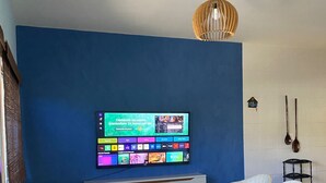 A living room with a blue wall, a mounted TV, two white armchairs, a small coffee table, and a hanging light fixture. - Cozy house near the beach (Barra dos Coqueiros)