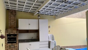 A patio area with a white table and chairs, a brick fireplace, a kitchen with white cabinets, and a pool in the background. - Cozy house near the beach (Barra dos Coqueiros)