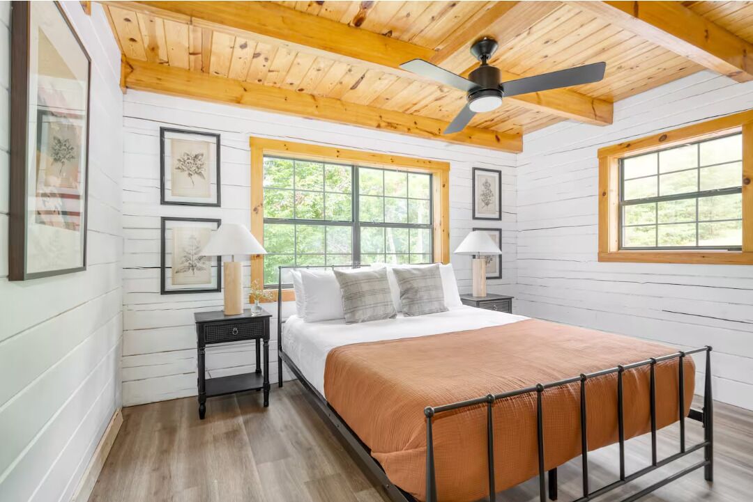 Une chambre à coucher avec un lit, un plafond en bois, deux fenêtres et des œuvres d’art encadrées accrochées aux murs.