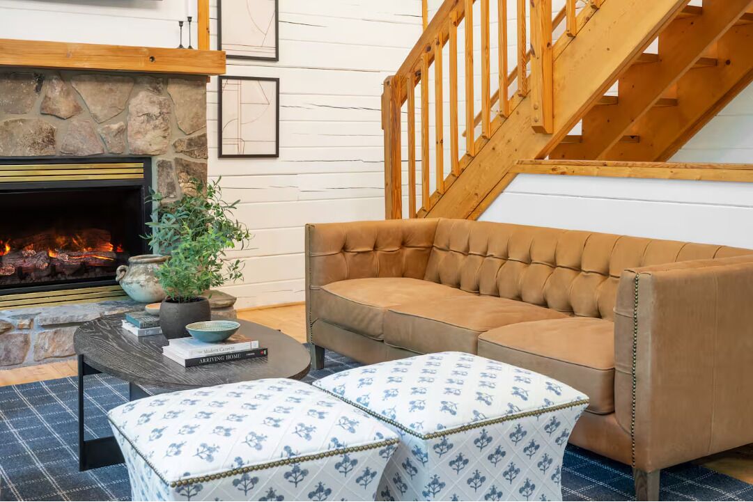Un salon chaleureux avec une cheminée en pierre, un canapé en cuir marron, un pouf à motifs et un escalier en bois.