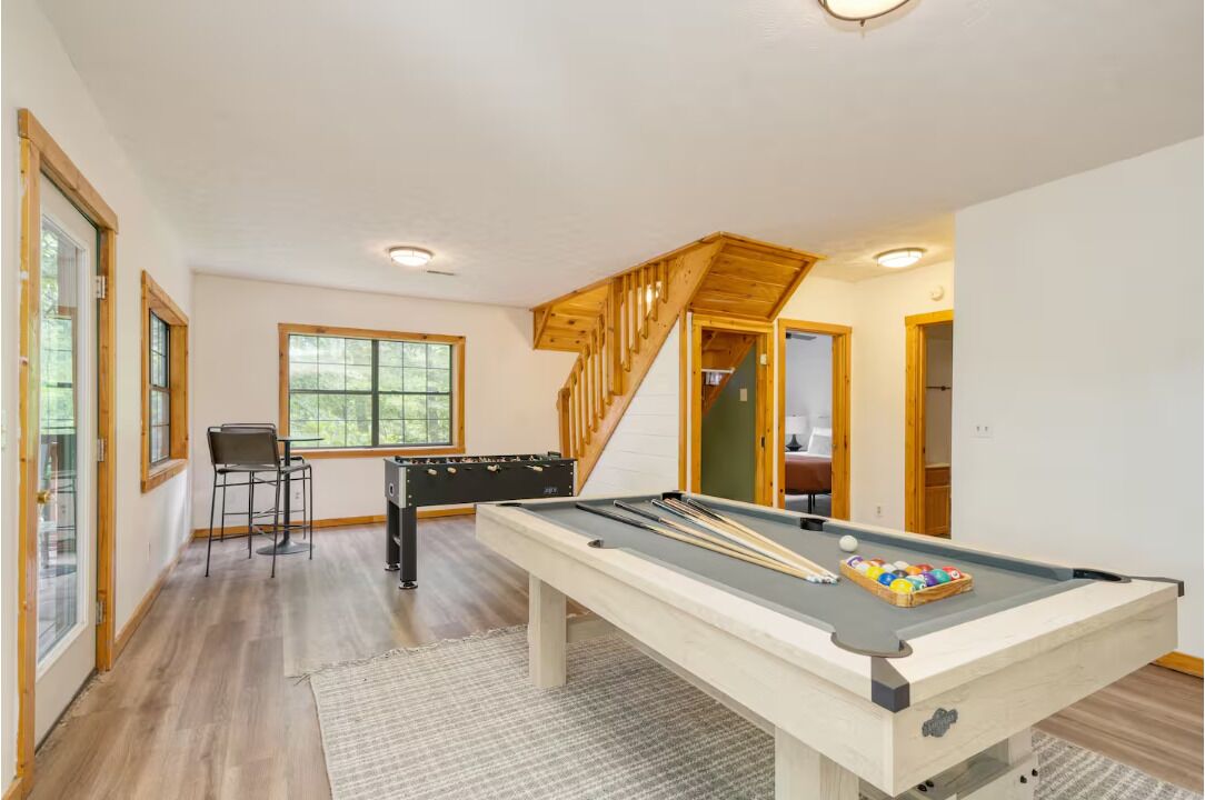 Een pooltafel in een ruimte met een houten trap, een zithoek met stoelen en grote ramen.