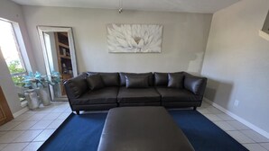 A living room with a sectional sofa, a matching ottoman, a large abstract painting, and a view of the outdoors through a door. - The Rancho Viejo Getaway (Rancho Viejo)