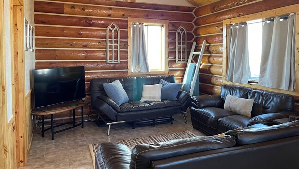 L’intérieur d’une cabane en rondins, avec un canapé en cuir noir, un téléviseur et une échelle appuyée contre le mur.