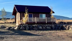 Une cabane en rondins dotée d’une terrasse en bois et de fondations en pierre, entourée d’un paysage aride et de montagnes.