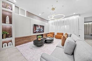 A modern living room with a grey sofa, two black round coffee tables, a TV mounted on the wall, and a fireplace. - 7BR house with private pool near Disney (Kissimmee)