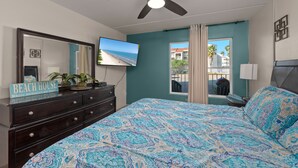 Un dormitorio con cama, ventilador de techo, televisor, cómoda, ventana con cortinas y un cartel que dice "BEACH HOUSE".