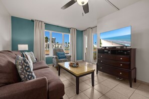 Een woonkamer met turquoise muren, een bruine bank, een houten salontafel, een tv op een meubel en uitzicht op het strand door grote ramen.