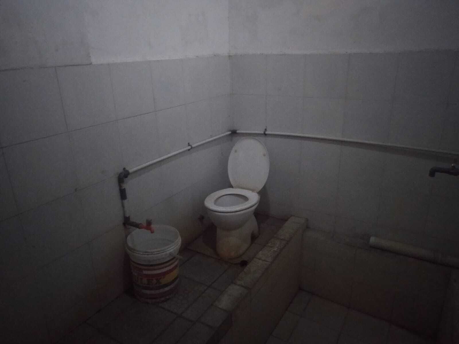 A small, dimly lit bathroom with a toilet, a bucket, and a shower area.