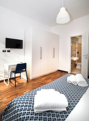Une chambre moderne avec un téléviseur à écran plat fixé au mur, un bureau avec une chaise, un lit recouvert d’une couvre-lit à motifs et une salle de bain visible à travers une porte ouverte.