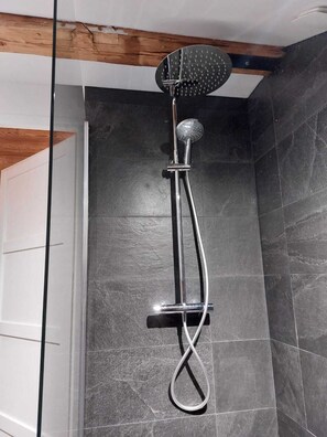 Eine Dusche mit Handbrause und Deckenbrause, vor einer dunkel gefliesten Wand mit einem Holzbalken an der Decke.