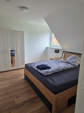 Quarto com teto inclinado, cama com colchão escuro e roupa de cama floral, guarda-roupa de madeira e uma pequena mesa de cabeceira com abajur.