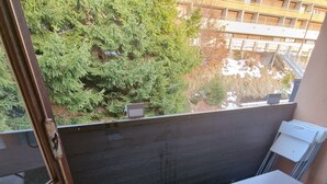 En balkong med konstgräs, ett litet bord och ett fönster med utsikt över träd och en byggnad.