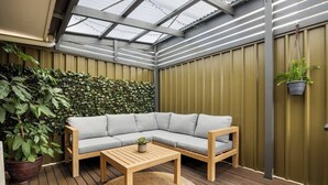 Ein moderner Außenbereich mit Holzdeck, einem grauen Ecksofa, einem Holztisch und einem großen Topfpflanzen.