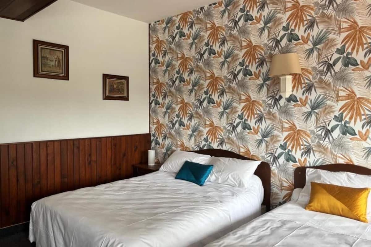 Habitación de hotel con dos camas, cabecera de madera, papel tapiz floral, cuadros en la pared y una lámpara de noche.