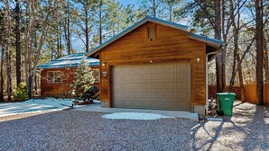 A wooden cabin with a garage, surrounded by trees and a gravel driveway. - Fire Pit + Yard: Woodsy Retreat Near Woodland Lake (Pinetop)