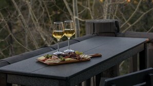 A table with a charcuterie board and two glasses of white wine on a balcony overlooking a mountainous landscape. - Luxe Mtn Cabin | Hot Tub, Sauna, Stargazing Dome (Bailey)