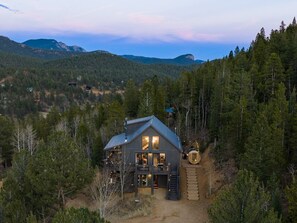 A cabin with a large porch, a staircase leading to the entrance, and a view of the surrounding forest and mountains. - Luxe Mtn Cabin | Hot Tub, Sauna, Stargazing Dome (Bailey)