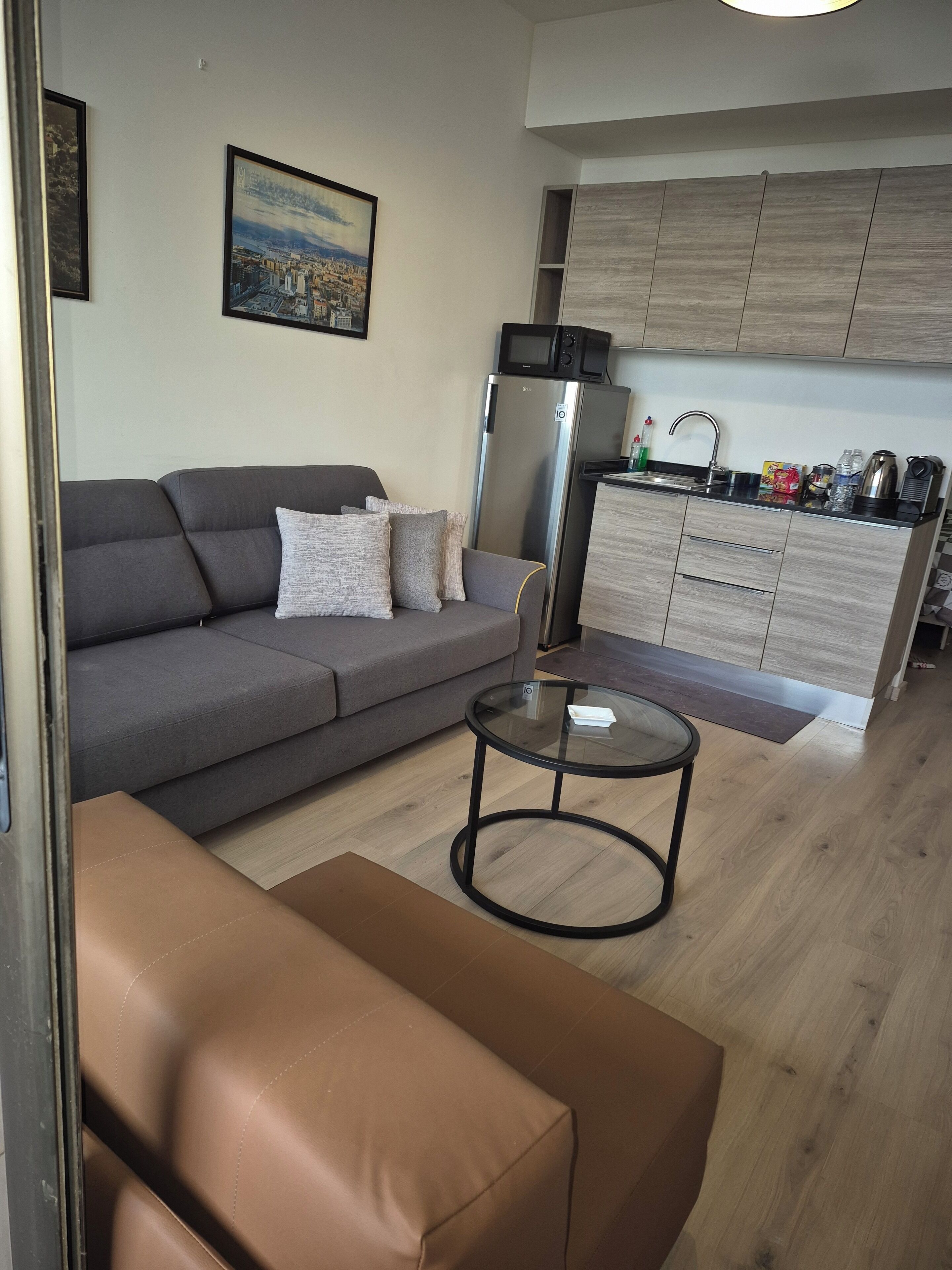 A modern living room with a grey sofa, a glass coffee table, and a brown ottoman. A kitchen area with wooden cabinets and a microwave is visible in the background.