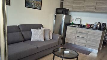 A modern living room with a grey sofa, a glass coffee table, and a brown ottoman. A kitchen area with wooden cabinets and a microwave is visible in the background.