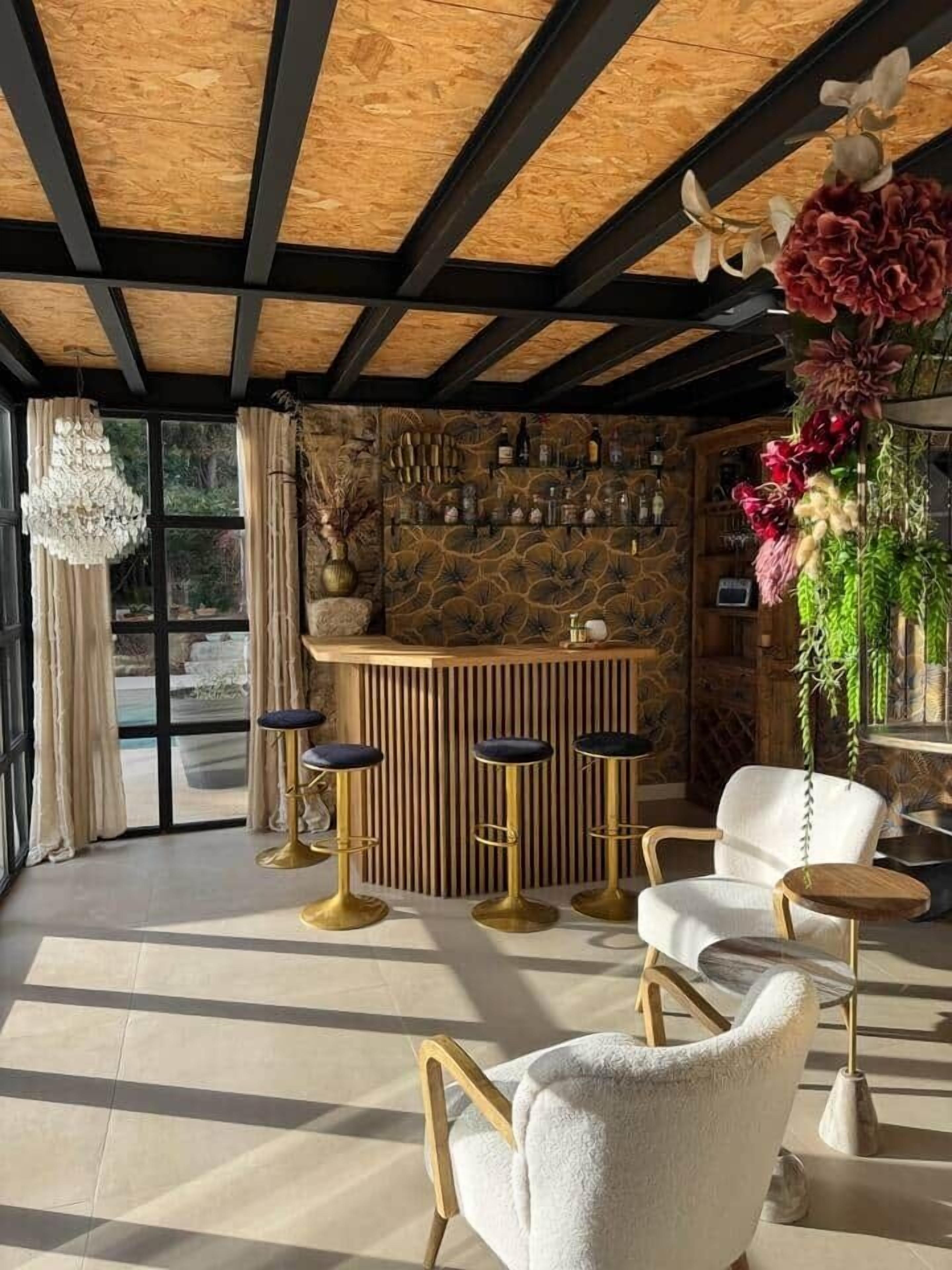 Un espace bar avec un plafond en bois, un mur en pierre et des tabourets de bar. On y trouve également des chaises et une plante suspendue.