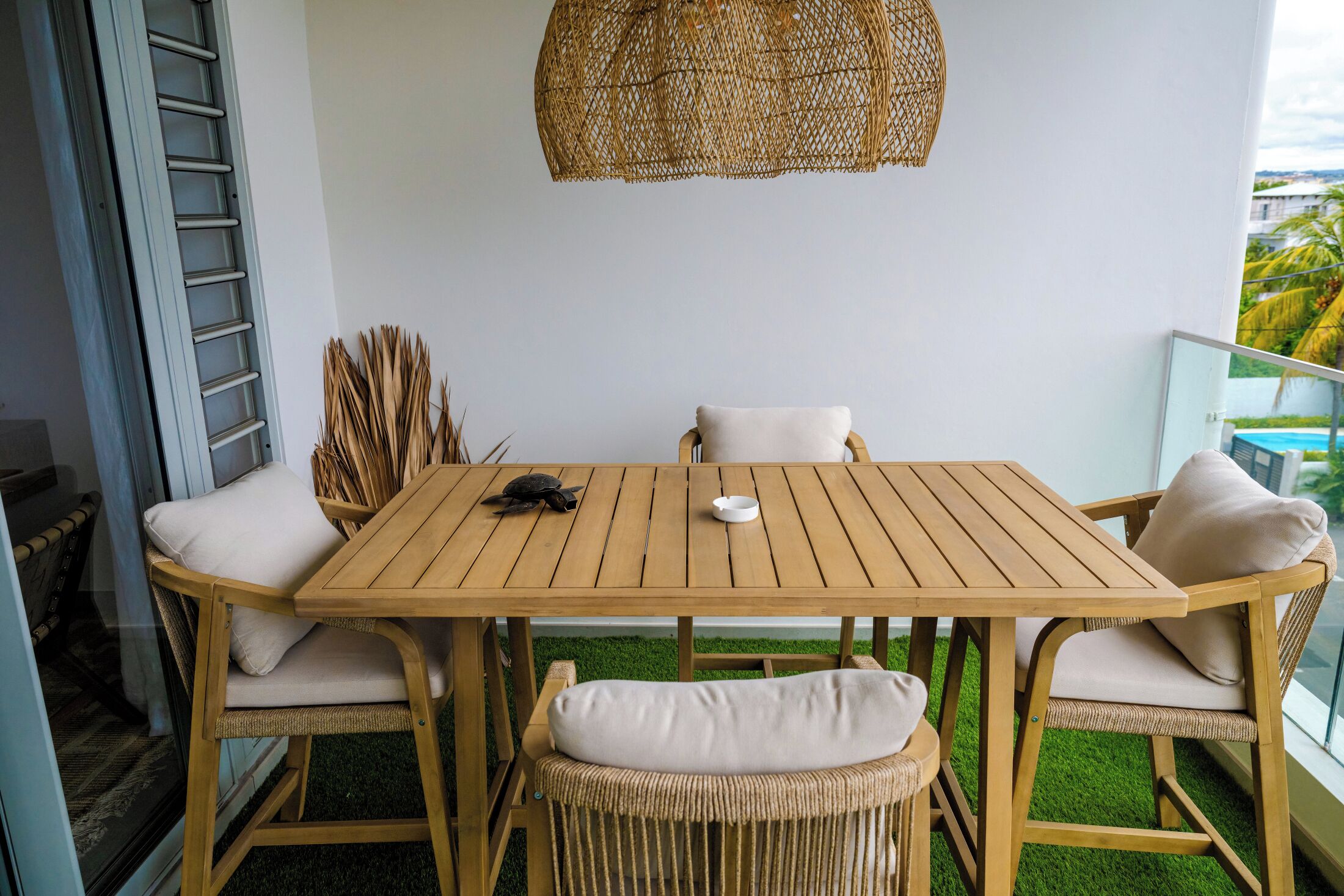 Een houten tafel met witte kussens, een geweven hanglamp en uitzicht op de stad vanaf een balkon.