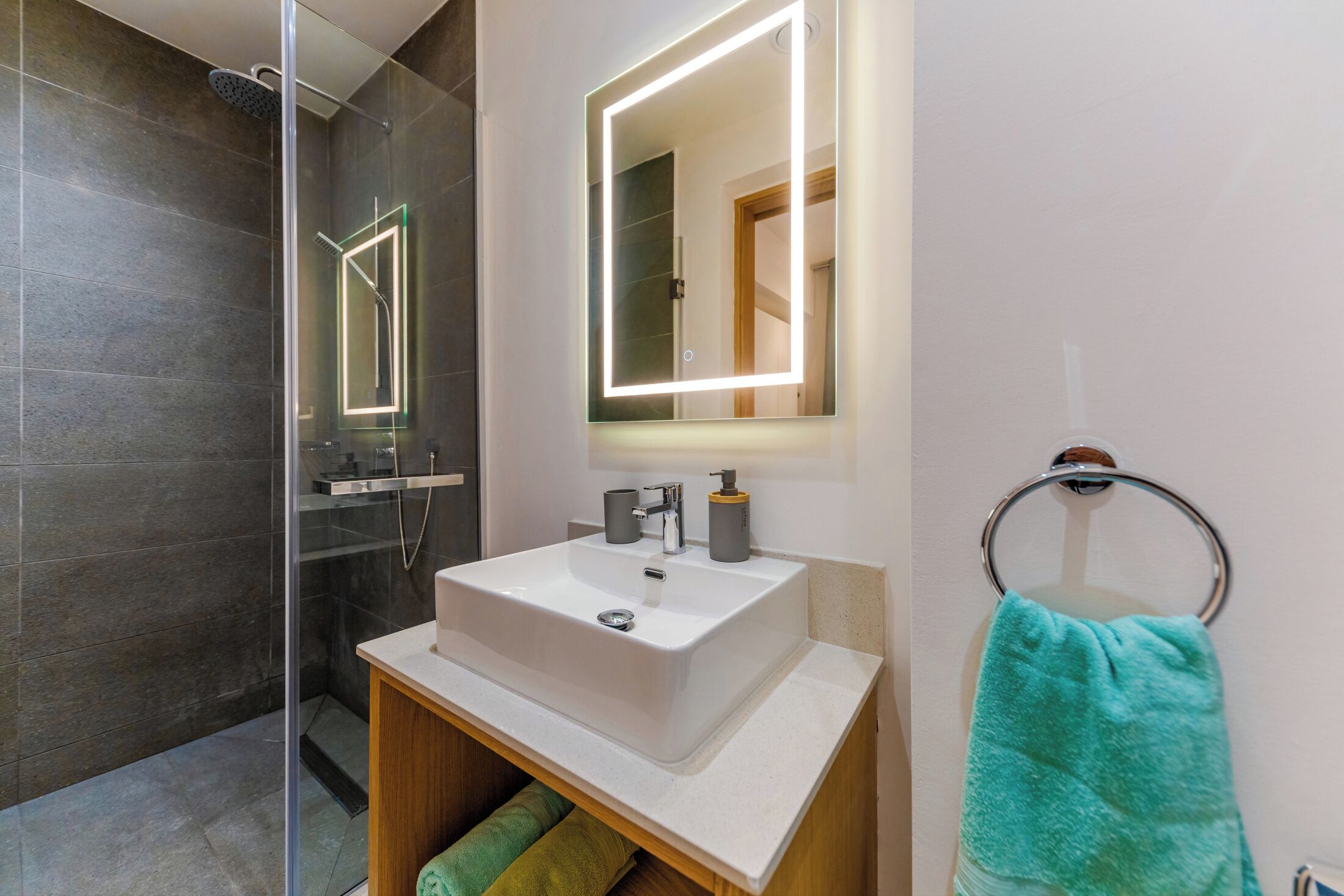 Une salle de bain moderne avec une douche à parois de verre, un lavabo blanc avec un miroir au-dessus et un porte-serviettes orné d’une serviette verte.