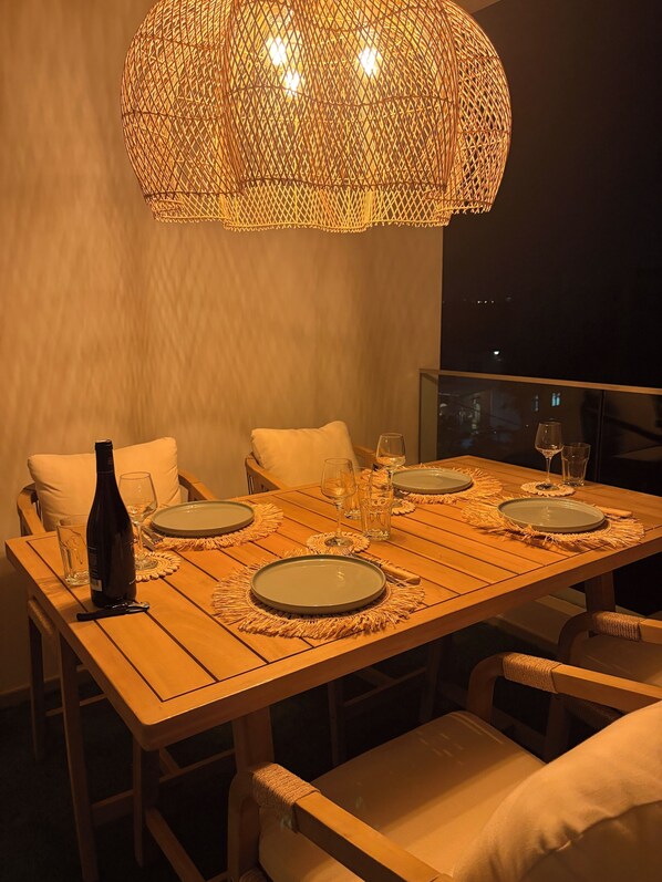 A dining table set for four with plates, glasses, and a bottle, surrounded by chairs with cushions.