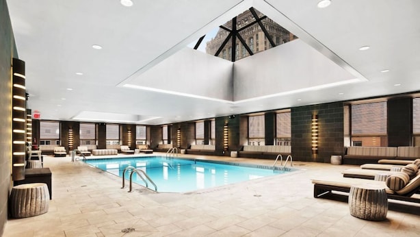 Un espace piscine intérieur moderne, doté d’un plafond au design géométrique, de sièges disposés autour de la piscine et offrant une vue sur le paysage urbain grâce à une verrière.