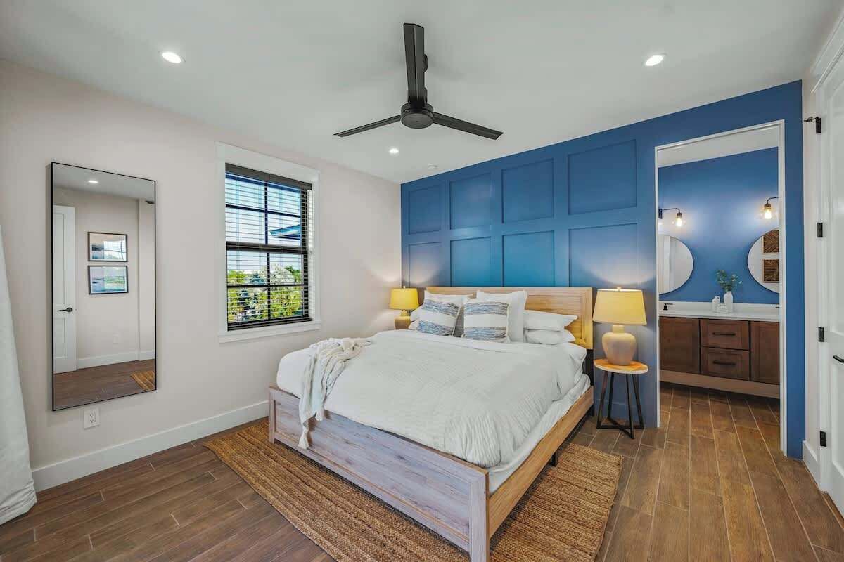 A bedroom with a bed, a ceiling fan, a window, a mirror, and a bathroom with a sink and mirror.
