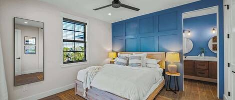 A bedroom with a bed, a ceiling fan, a window, a mirror, and a bathroom with a sink and mirror.